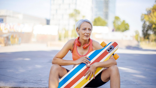 Ältere sitzende Frau mit sportlicher Bekleidung, schnurlosen Kopfhöhrern und einem Longboard in der Hand